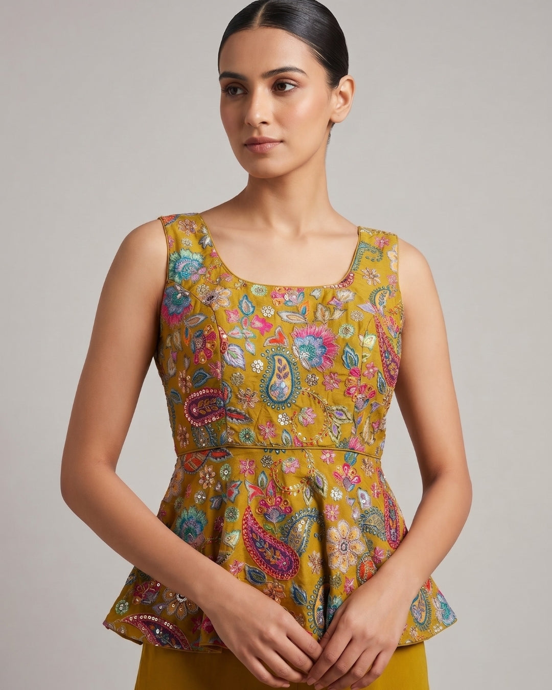A woman wearing a sleeveless mustard yellow peplum top with a colorful paisley and floral print, paired with matching yellow bottoms, photographed against a neutral gray studio background.