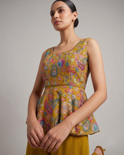 A woman wearing a sleeveless peplum top with a vibrant multicolor paisley and floral print on a mustard yellow base, paired with matching yellow trousers, photographed against a neutral gray studio background.