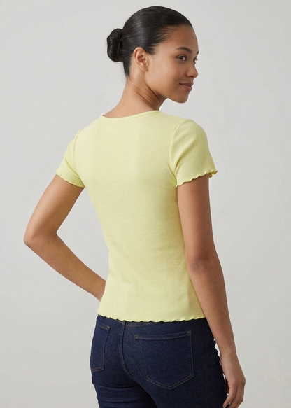 A woman shown from the back wearing a pale yellow short-sleeve top with scalloped edges and dark blue jeans, standing against a plain light gray wall.